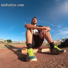 A person wearing a black athletic outfit is seated on a wooden platform in an outdoor sports field. The individual is relaxed, with arms resting on knees. The field is expansive with a clear blue sky and scattered clouds above. A brightly colored running shoe and athletic gear are visible, along with a water bottle and a blue backpack placed beside the person. Lampposts and residential buildings can be seen in the background.