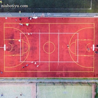 An aerial view of a sports court with a vibrant red surface and yellow lines marking different sections for basketball and soccer. Several people are present on the court, some engaged in activities, while others are standing along the perimeter. The court is enclosed by a metal fence and surrounded by grass on all sides.