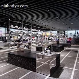 A modern sneaker store with sleek black shelving displaying various athletic shoes and sports apparel. Central seating areas and spotlights provide a contemporary retail ambiance. The signage prominently features a famous sportswear slogan in the background.