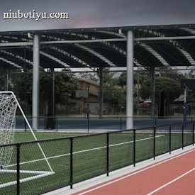 An outdoor sports facility featuring a running track and a soccer goal. The track is red, surrounded by a black metal fence, and the field is covered with artificial green turf. In the background, a basketball court is visible under a covered roof with metal supports. The sky is overcast, adding a moody atmosphere.