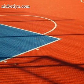 A vibrant sports court with sections of red and blue, featuring crisp white lines marking the boundaries. Shadows from a structure create geometric patterns across the surface, adding depth and interest to the scene.